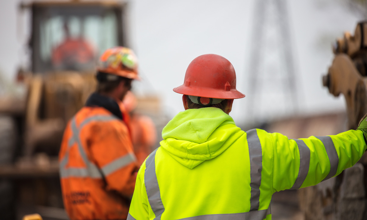 A construction worker wearing a hard hat and high vis jumper stands with his back towards the camera while on a construction site.